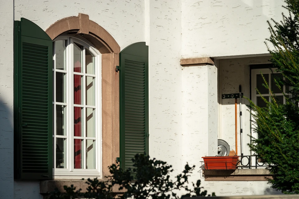 Un artisan menuisier en tenue professionnelle installant un volet battant sur la façade extérieure d'une maison résidentielle. L'artisan, équipé d'outils, est en train de fixer le volet battant à la fenêtre. La photo est prise de jour, montrant l'environnement extérieur de l'habitation, avec une nette mise au point sur le travail d'installation pour souligner le savoir-faire et la qualité d'exécution de l'équipe.