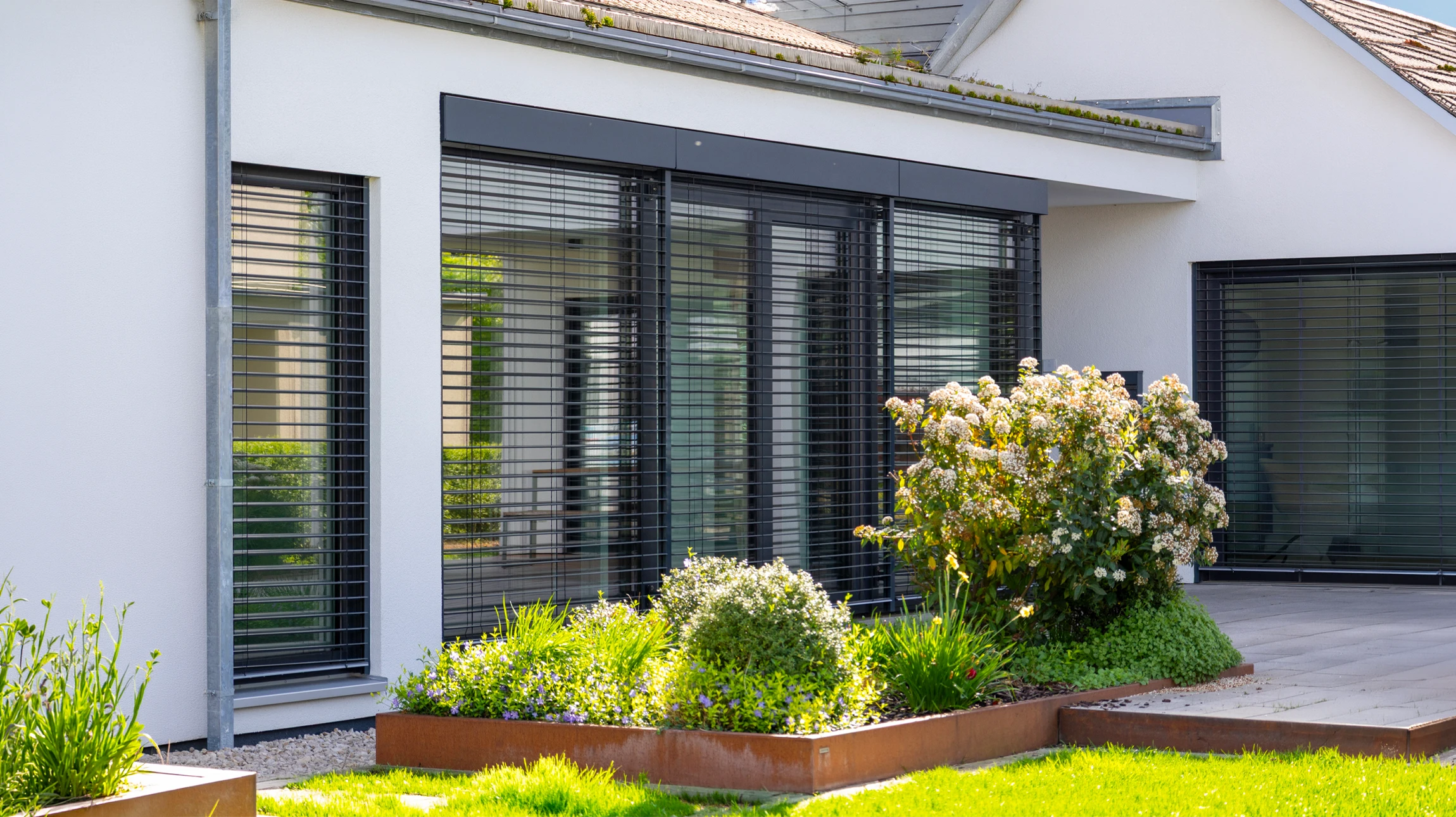 Façade de maison résidentielle moderne équipée de brise-soleil orientables en aluminium de couleur anthracite fixés horizontalement devant une large baie vitrée. Les lames sont partiellement ouvertes, laissant passer la lumière naturelle tout en créant de l'ombre sur la façade. Ambiance lumineuse diurne ensoleillée montrant le contraste entre les zones ombragées et éclairées. Architecture contemporaine avec revêtement clair, mise en avant de la qualité et de la fonctionnalité du produit installé.